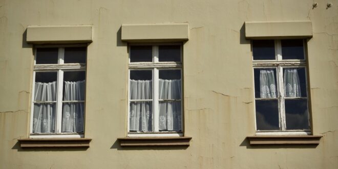 Drei Fenster mit weißen Rahmen und Gardinen in einer hellen, rissigen Altbaufassade