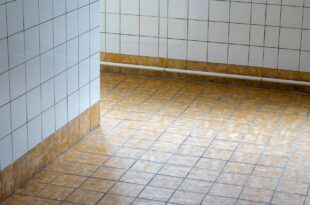 Gefliester Boden und geflieste Wand in einem Bad, am Rand Sockelfliesen und ein Rohr an der Wand