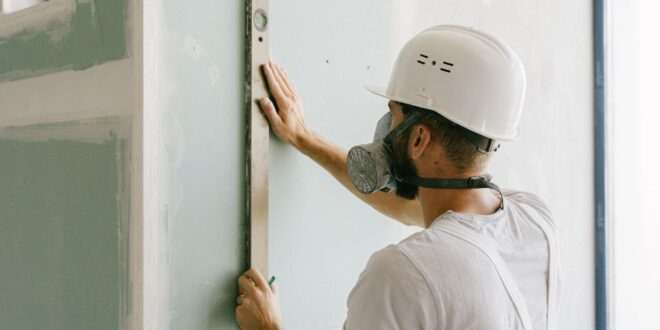 Handwerker mit Atemschutz und Wasserwaage an einer grünen Gipskartonwand