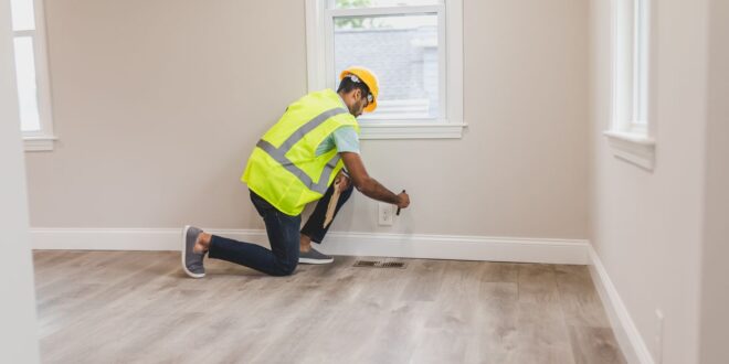 Arbeiter mit Warnweste kniet in einem leeren Raum mit Holzboden und prüft an der Wand etwas auf Bodenhöhe