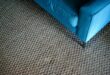 Grob gewebter, grau-brauner Teppichboden in Berber-Optik mit der Ecke eines blauen Sofas
