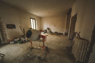 Entkernter Raum mit rauem Bodenuntergrund, Betonmischer, Farbeimern und einem alten Heizkörper während der Renovierung